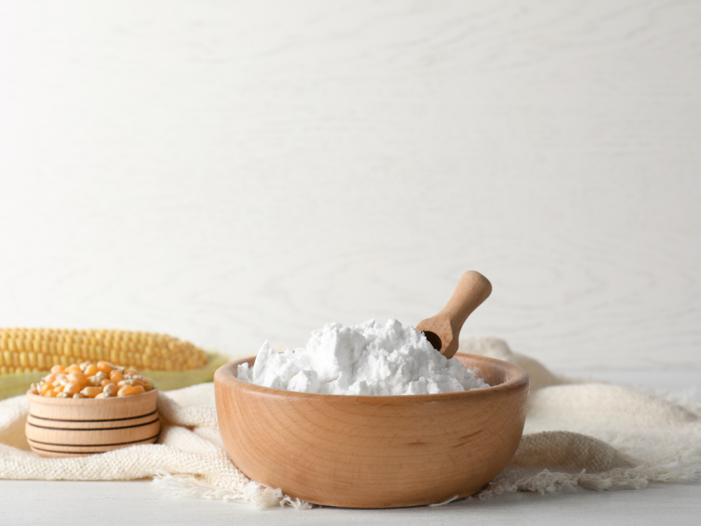wooden bowl of cornstarch with wooden scooper. cloth underneath and a smaller bowl with cork kernels. An ear of corn in the background